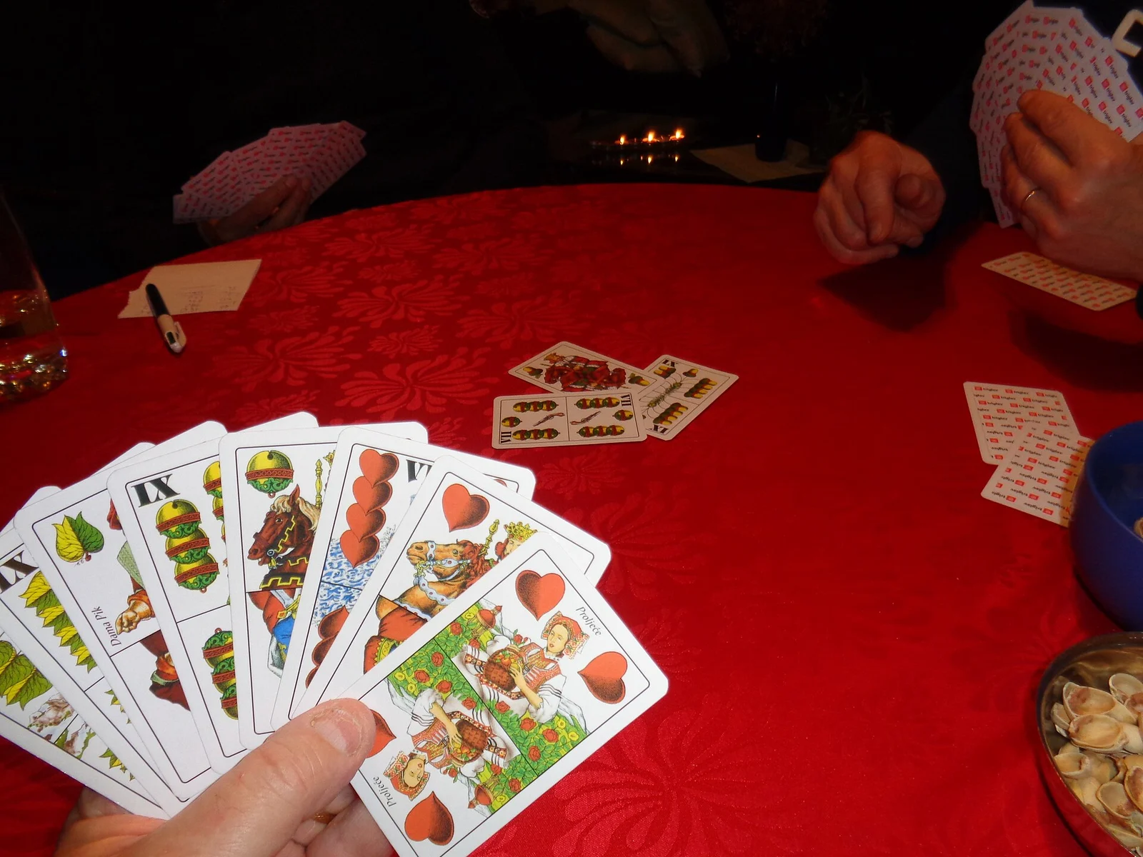 Kartenfächer in der Hand eines Spielers, ausgespielte Karten auf rotem Tischtuch.