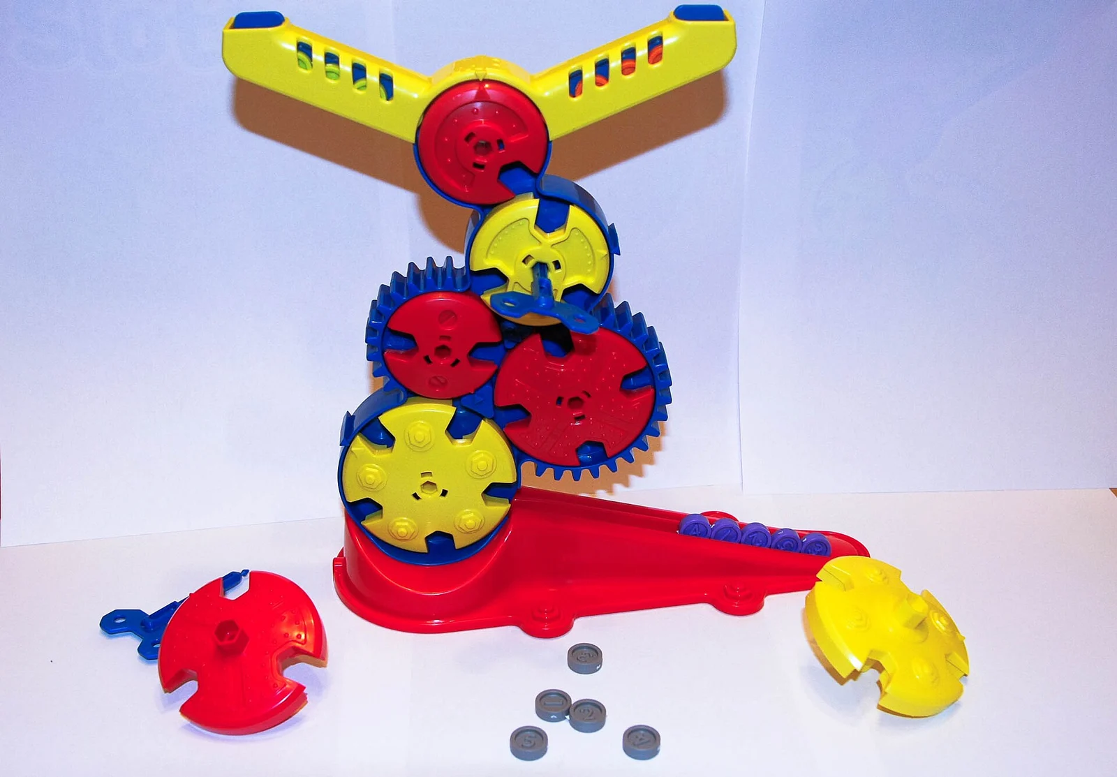Aufgebautes Geschicklichkeitsspiel aus rot-gelb-blauem Kunststoff mit gestapelten Zahnrädern und grauen Spielchips vor neutralem Hintergrund.