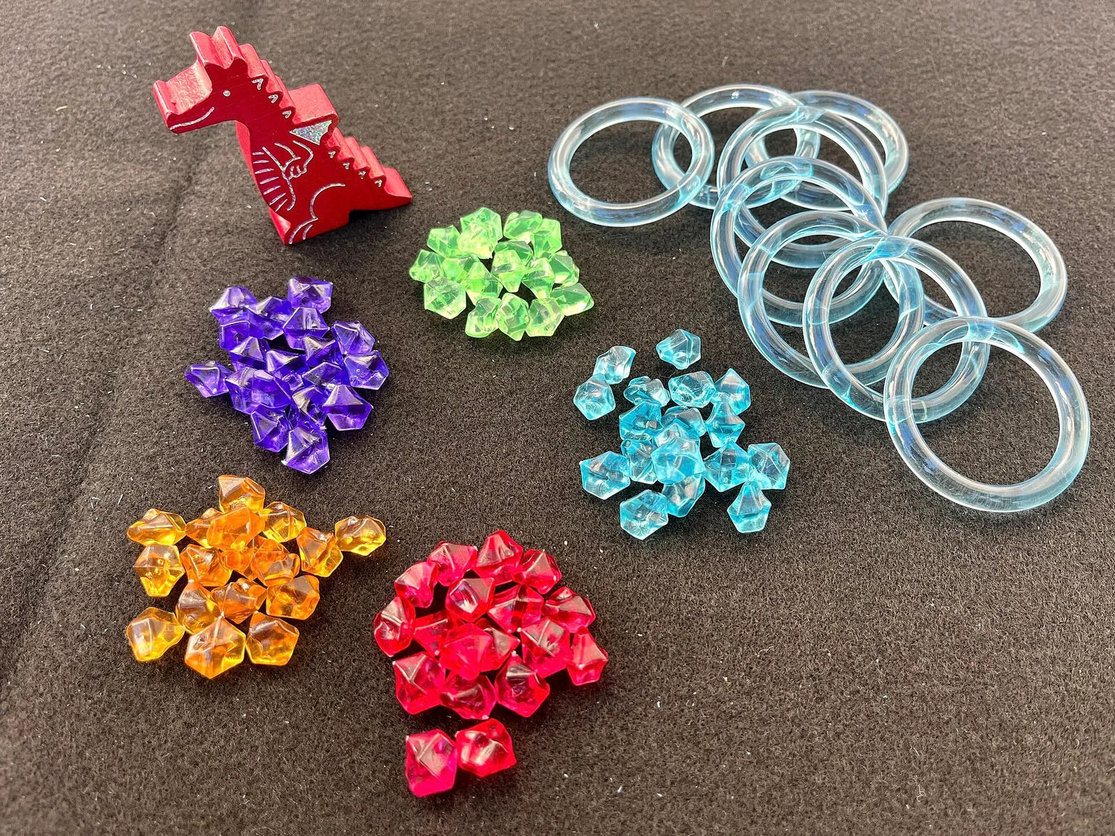 Spielmaterial: roter Holzdrache, durchsichtige blaue Ringe und nach Farben sortierte Funkelsteine auf grauem Untergrund.