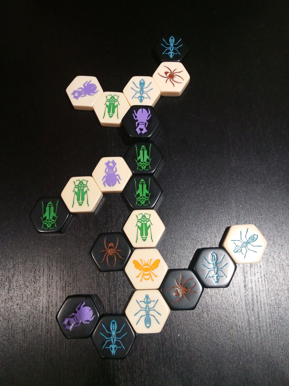 Sechseckige Hive-Spielsteine in Schwarz und Beige mit Insektenmotiven bilden auf dunklem Tisch einen Schwarm.