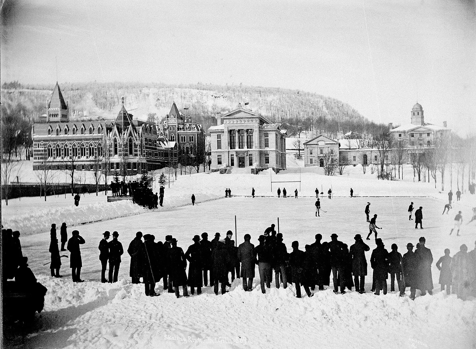 Historische Schwarzweißfotografie eines Hockeyspiels auf einer Freiluft-Eisfläche mit Zuschauern vor Universitätsgebäuden.