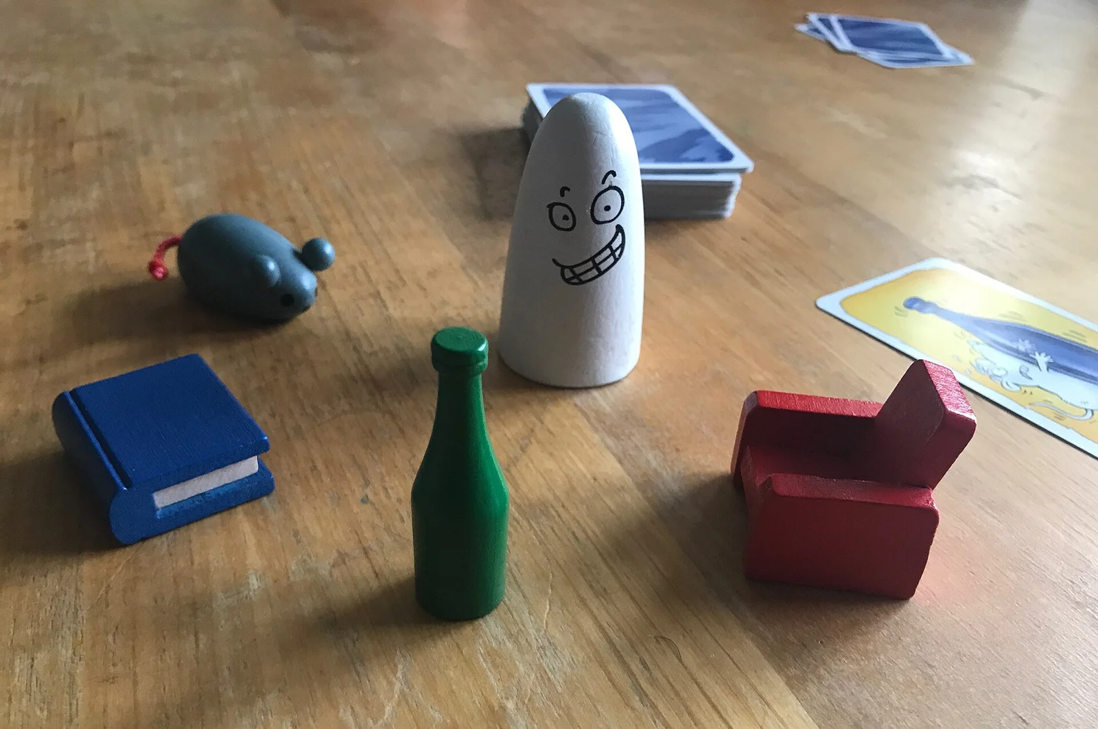 Fünf Holzfiguren – weißer Geist, grüne Flasche, blaues Buch, roter Sessel und graue Maus – auf einem Holztisch neben Kartenstapeln.