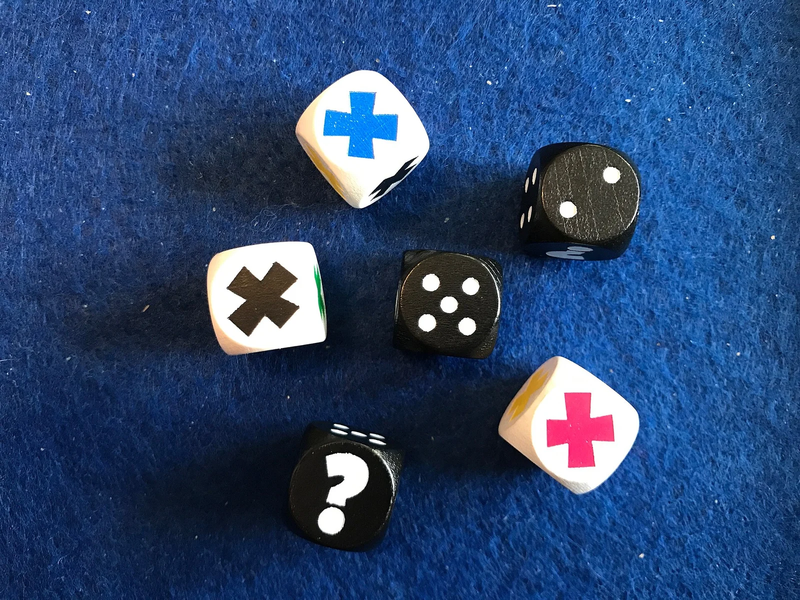 Sechs Würfel mit Farbkreuzen, Augenzahlen und Fragezeichen liegen auf blauem Filz.