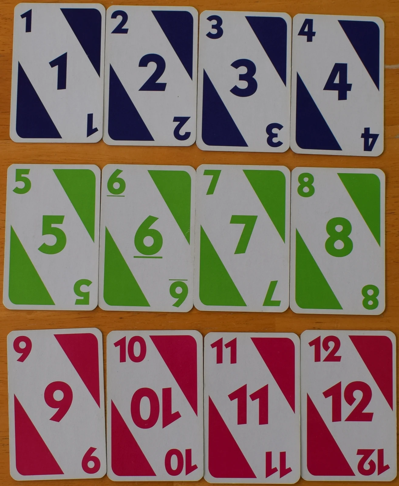 Zwölf nummerierte Spielkarten von 1 bis 12 in Blau, Grün und Pink auf einem Holztisch.