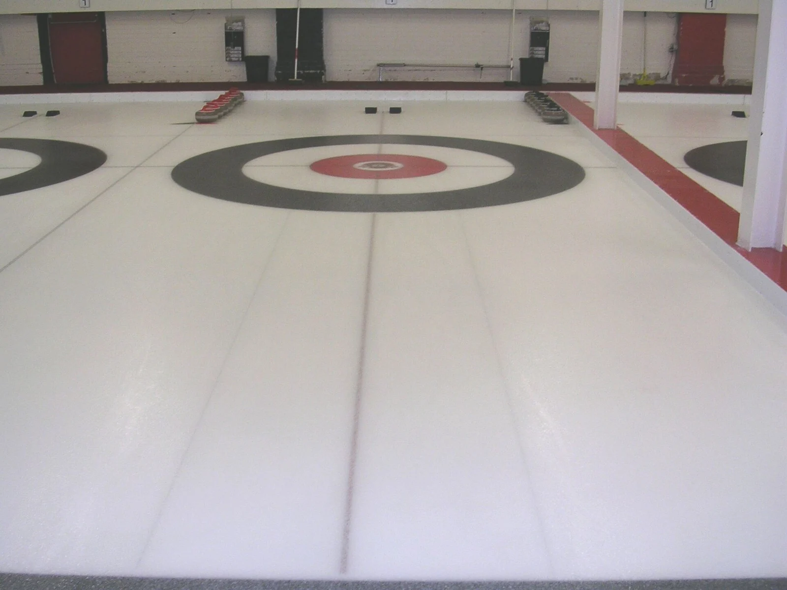 Curlingbahn mit Zielkreisen und am Rand abgestellten Steinen.