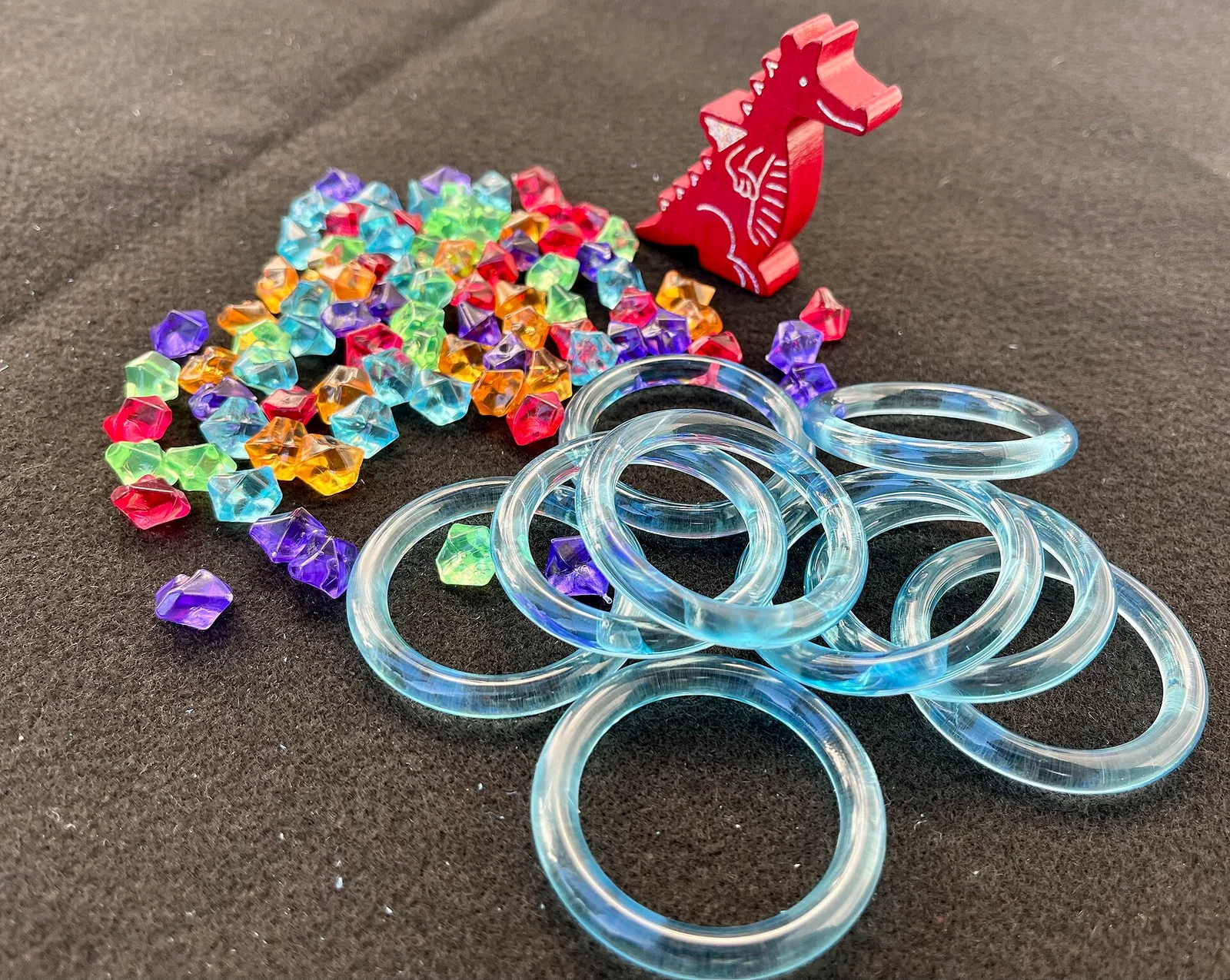 Bunte Funkelsteine, roter Holzdrache und ein Stapel durchsichtiger blauer Ringe auf grauem Untergrund.