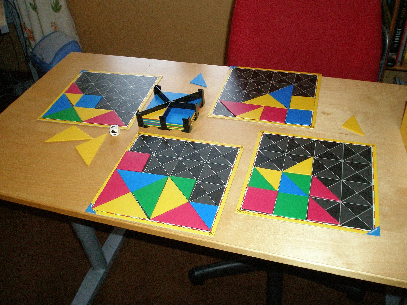 Vier Spieltafeln mit bunten Dreiecks- und Vierecksteilen auf einem Holztisch, dazu Würfel und Kartenhalter.