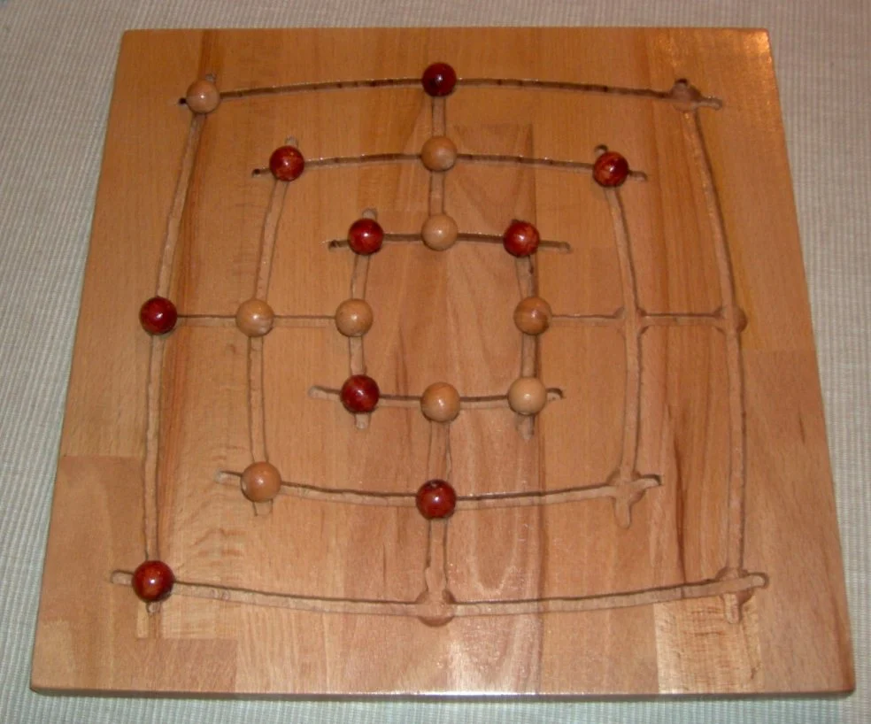 Hölzernes Mühlespielbrett mit eingeritzten Linien und roten sowie hellen Kugeln als Spielsteinen.
