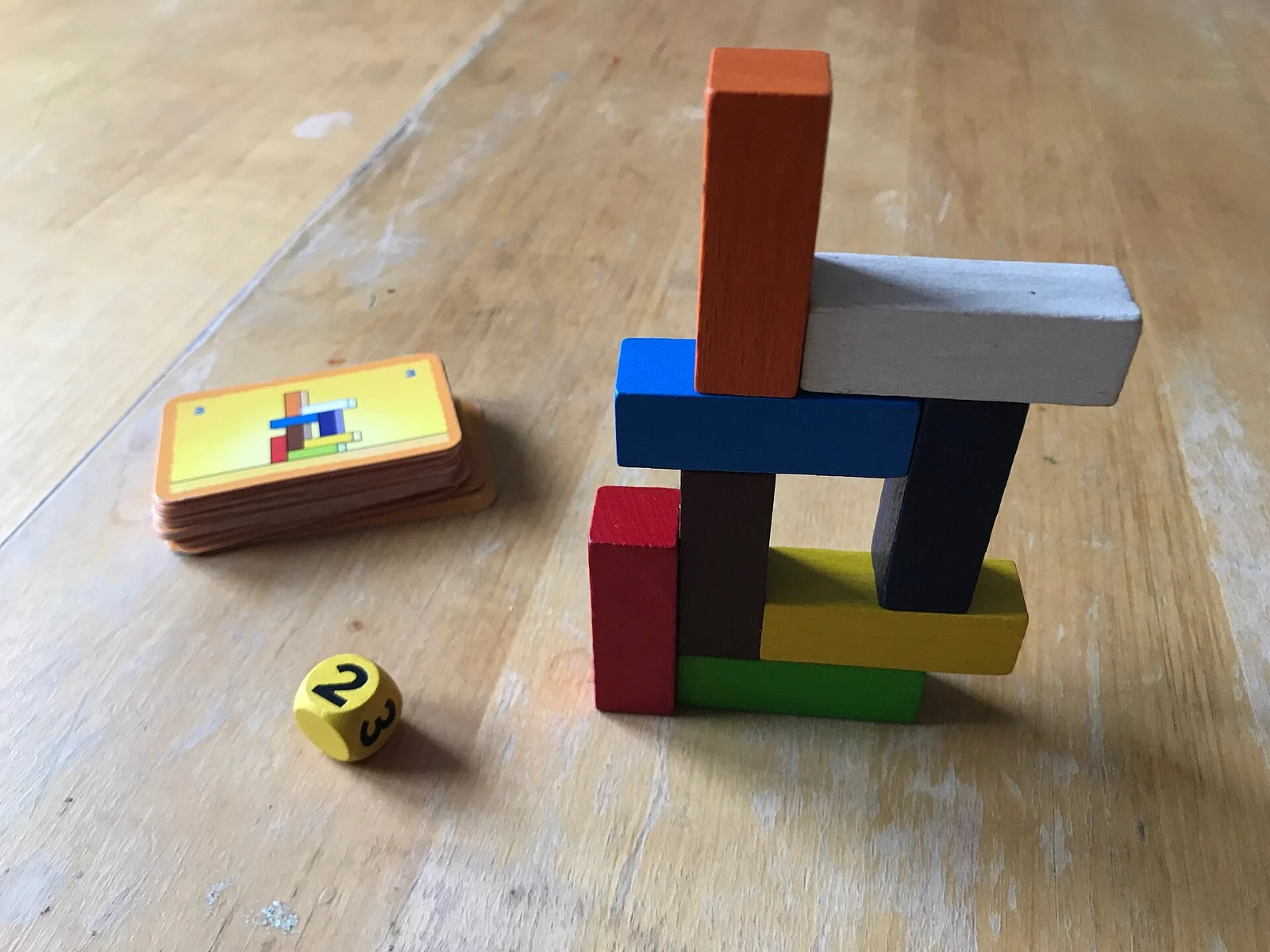 Bauwerk aus bunten Holzsteinen auf Holztisch, daneben ein Kartenstapel mit Bauvorlage und ein gelber Würfel.