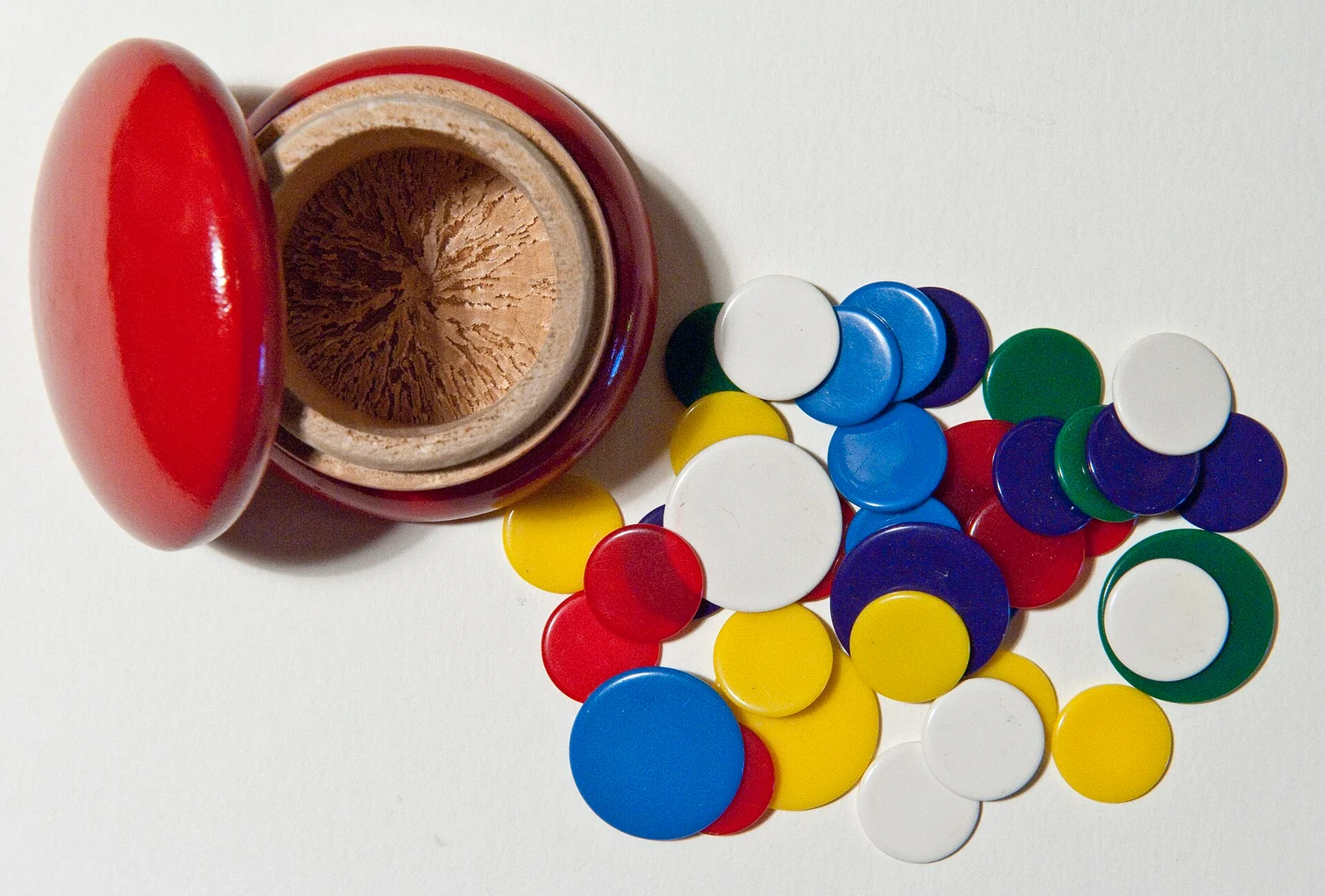 Roter Holzbecher mit abgenommenem Deckel und viele bunte Spielmarken in verschiedenen Größen auf hellem Untergrund.