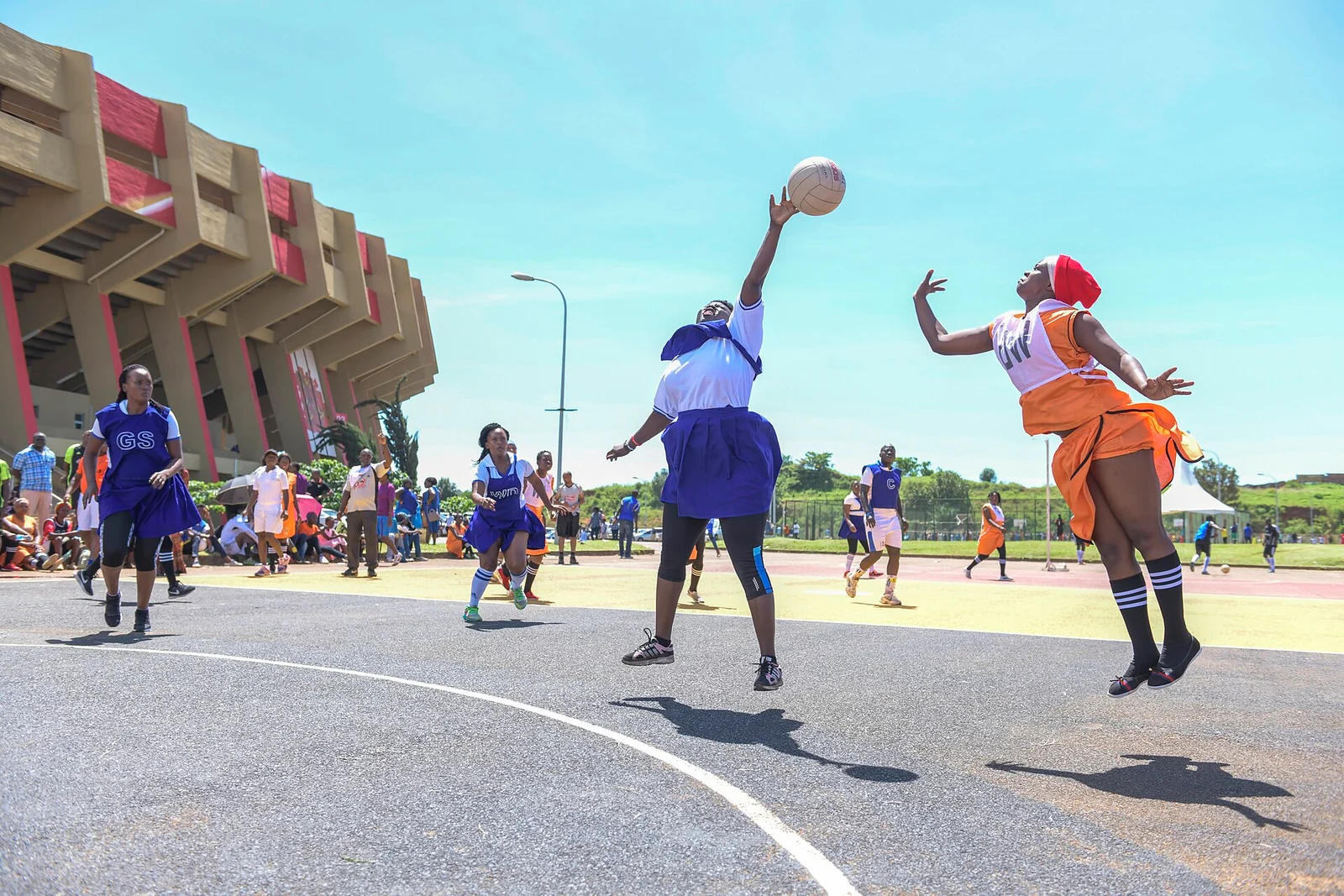 Zwei Netballspielerinnen springen im Zweikampf um den Ball auf einem Außenplatz vor einem Stadion.
