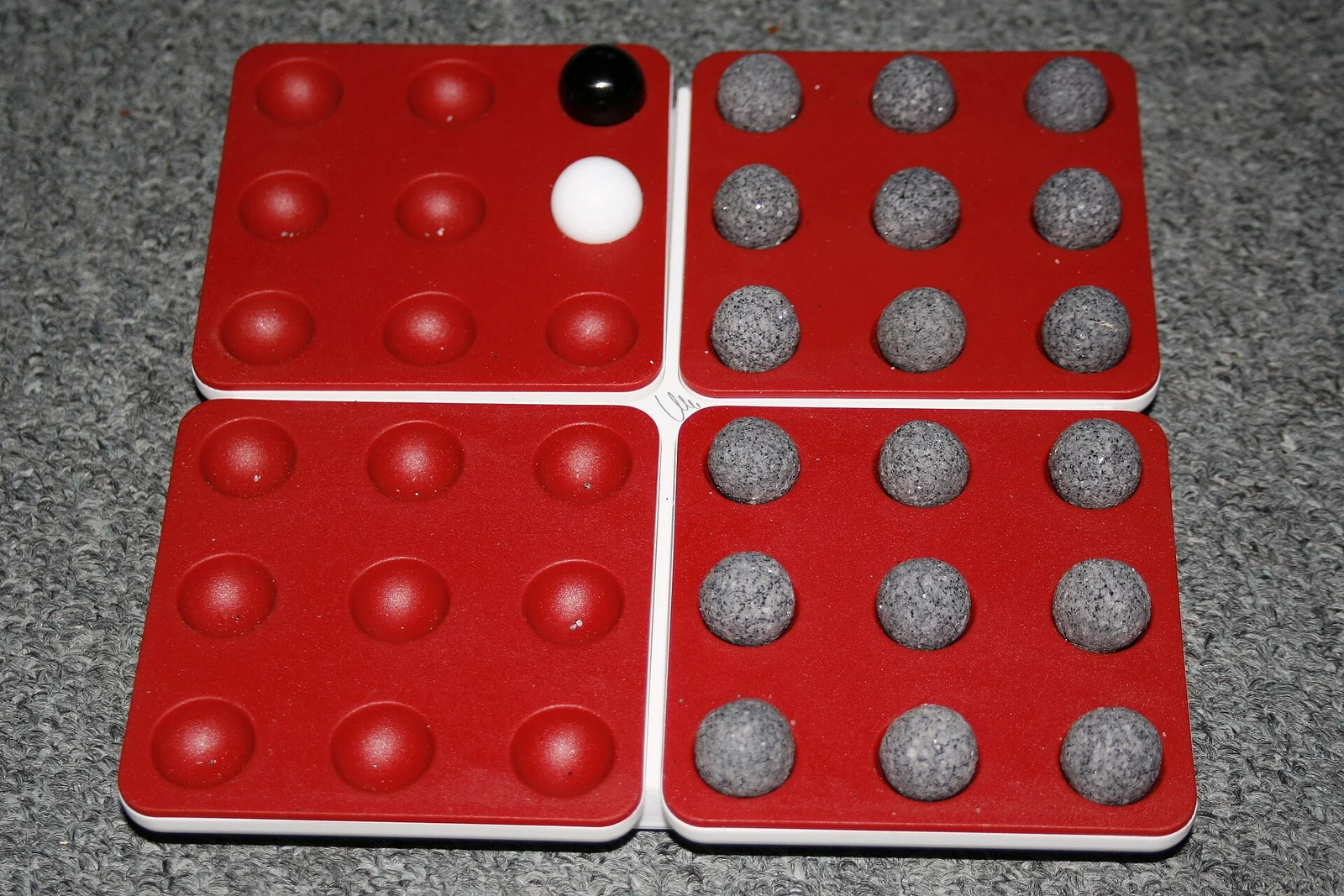 Rotes Pentago-Brett aus vier Teilquadraten mit Vertiefungen, teils mit grauen, schwarzen und weißen Kugeln belegt.