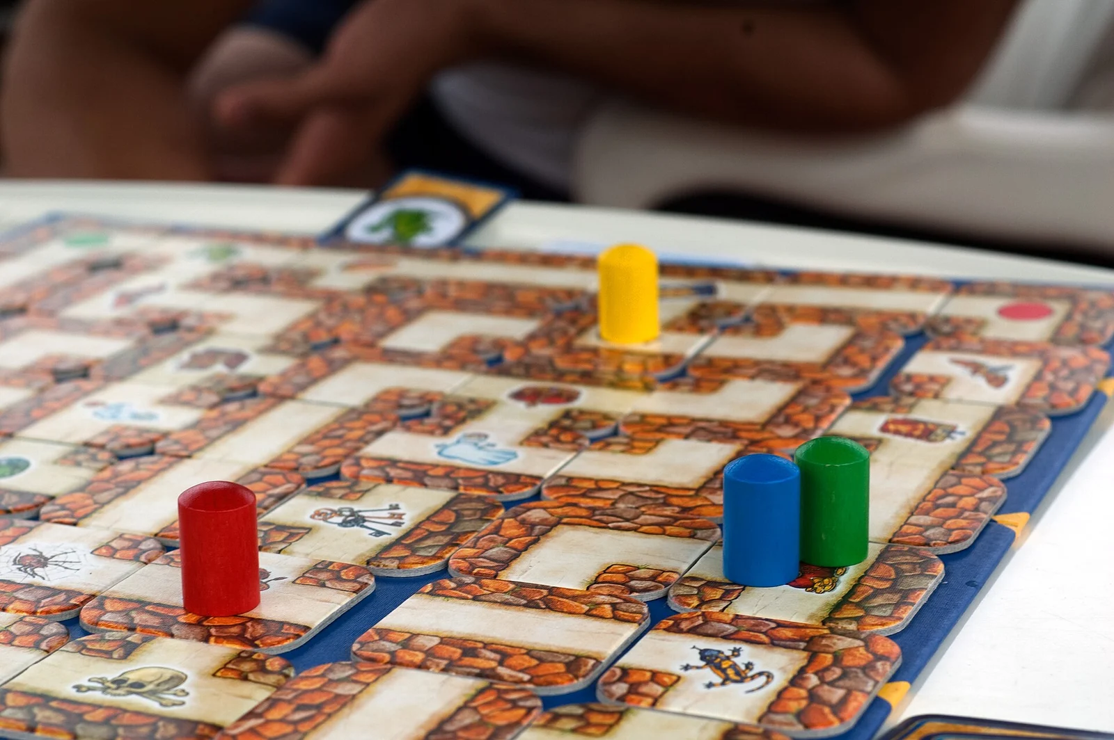Spielplan des verrückten Labyrinths mit verschiebbaren Gangkarten und vier farbigen Spielfiguren, im Hintergrund unscharf ein Mitspieler ohne erkennbares Gesicht.