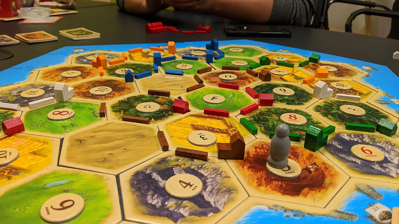 Catan-Spielplan während einer Partie mit Straßen, Siedlungen und Räuber, im Hintergrund ein Arm eines Mitspielers.
