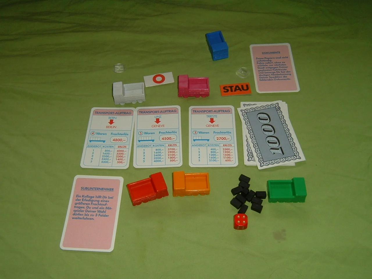 Spielmaterial von Auf Achse: bunte Lkw-Figuren, Transport-Auftragskarten, Ereigniskarten, Warenklötzchen und Würfel auf grünem Tuch.