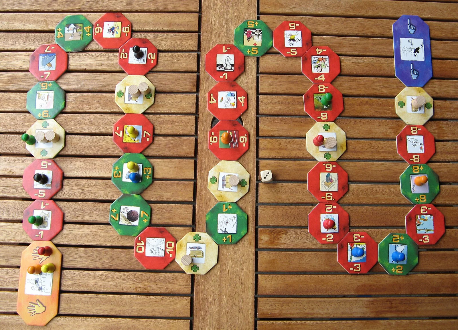 Ausgelegter Parcours aus achteckigen Plus-, Minus- und Glückstafeln mit Spielfiguren und einem Würfel auf einem Holztisch.