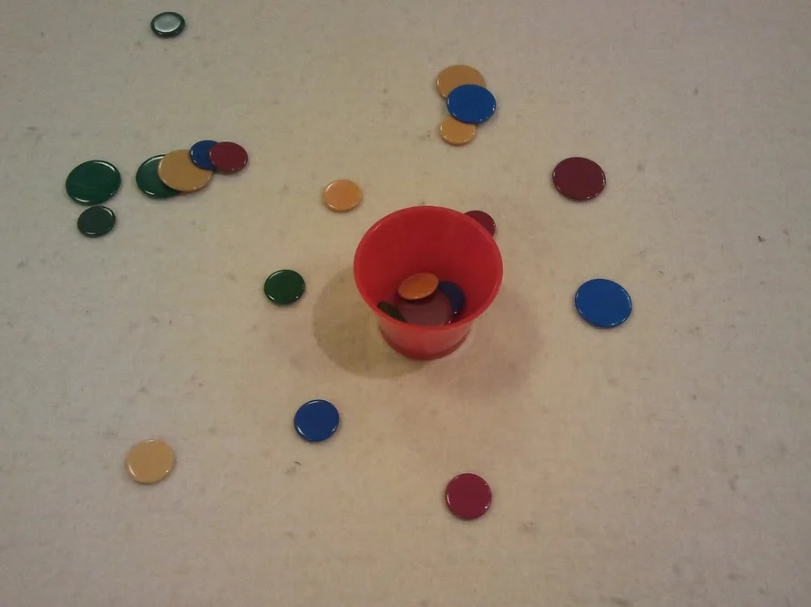 Roter Becher in der Tischmitte, umgeben von bunten Spielscheiben, einige liegen bereits im Becher.