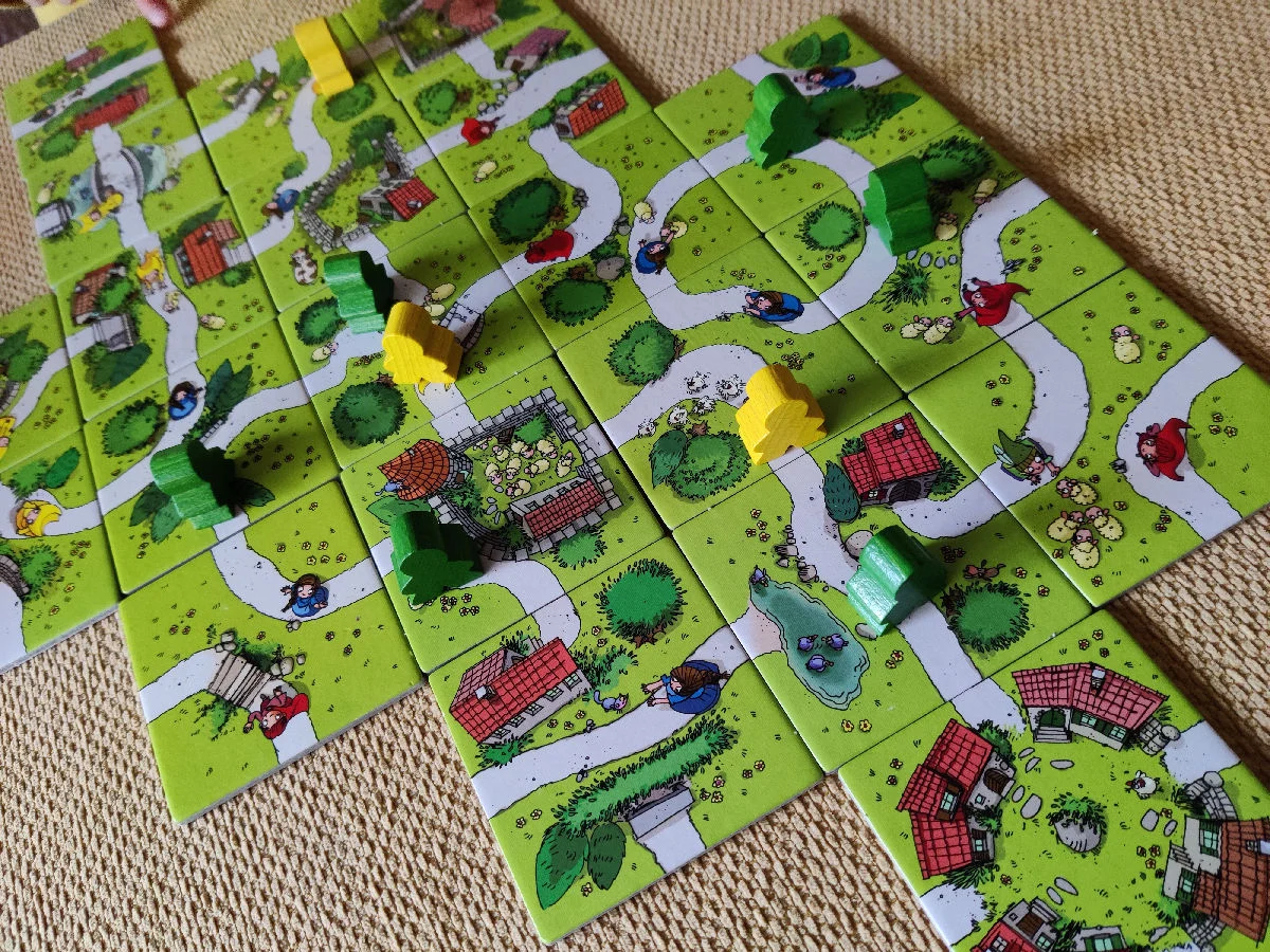 Ausgelegte Landschaftsplättchen mit Wegen, Häusern und bunten Holzfiguren auf einem Teppich.