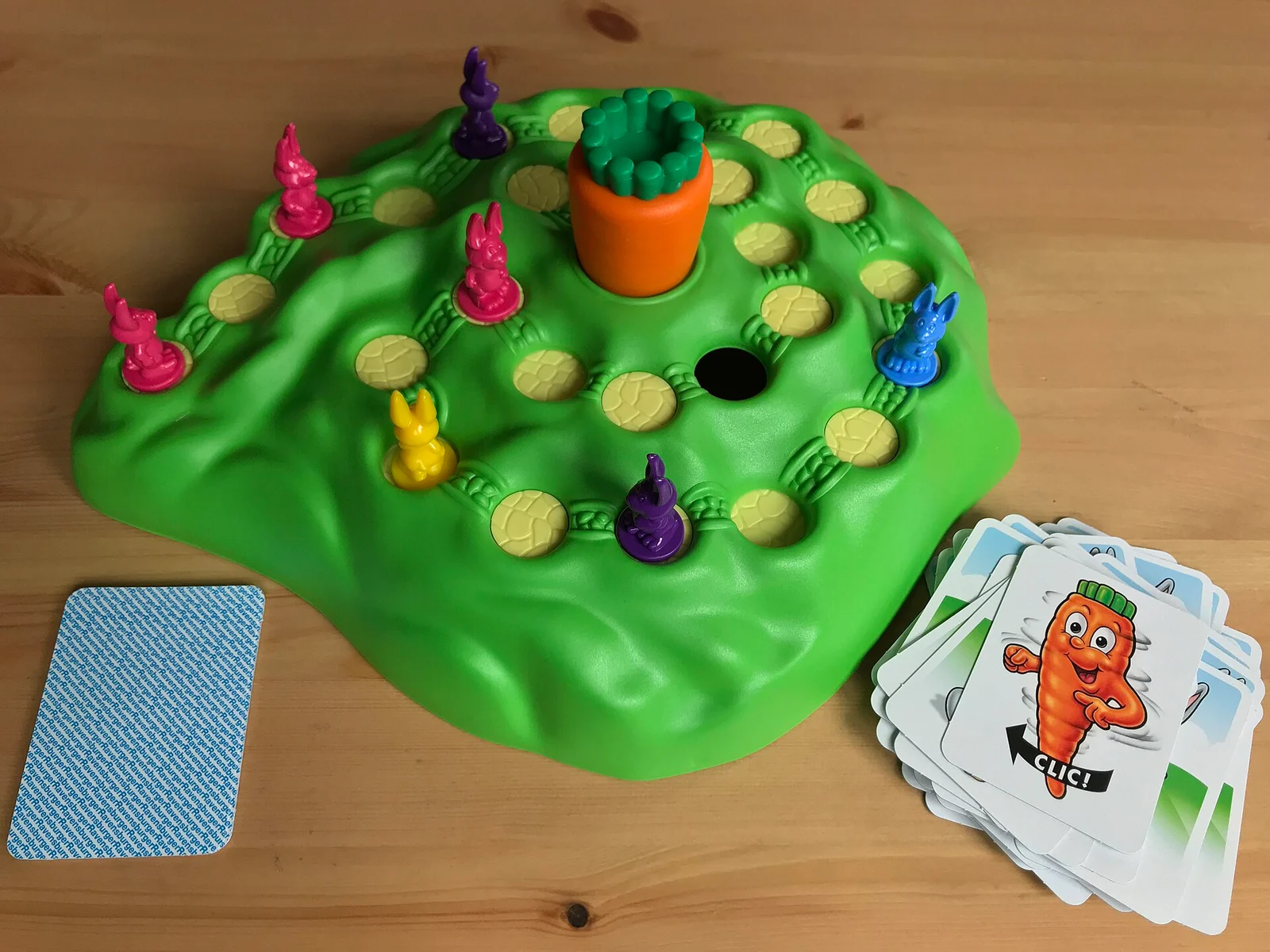 Grüner Spielhügel mit Löchern, bunten Hasenfiguren, Drehkarotte in der Mitte und Kartenstapeln auf einem Holztisch.