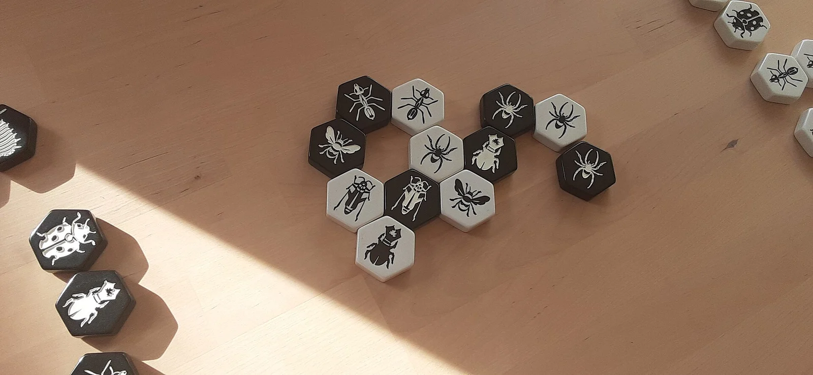 Schwarz-weiße sechseckige Spielsteine mit Insektensymbolen liegen auf einem Holztisch zu einem Schwarm zusammen.