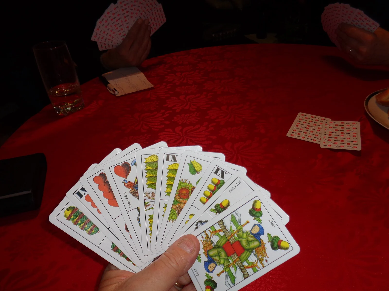Blick aus Spielerperspektive auf ein Kartenblatt über einem roten Tisch mit Mitspielern.