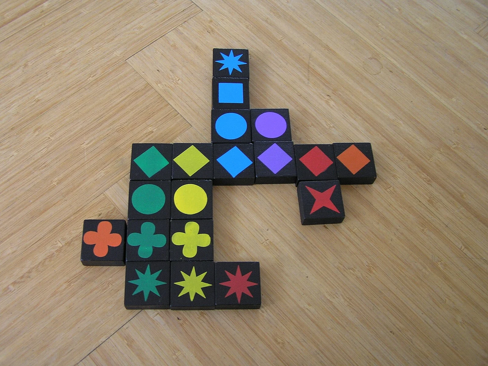 Auf einem Holzboden liegen schwarze Qwirkle-Steine mit farbigen Symbolen in gekreuzten Reihen.