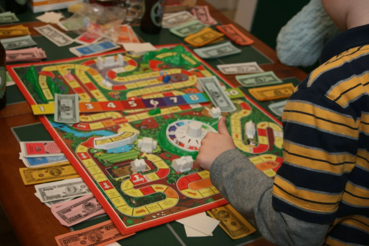 Person am Tisch dreht am Drehrad eines bunten Spielplans, umgeben von Spielgeld und Karten.
