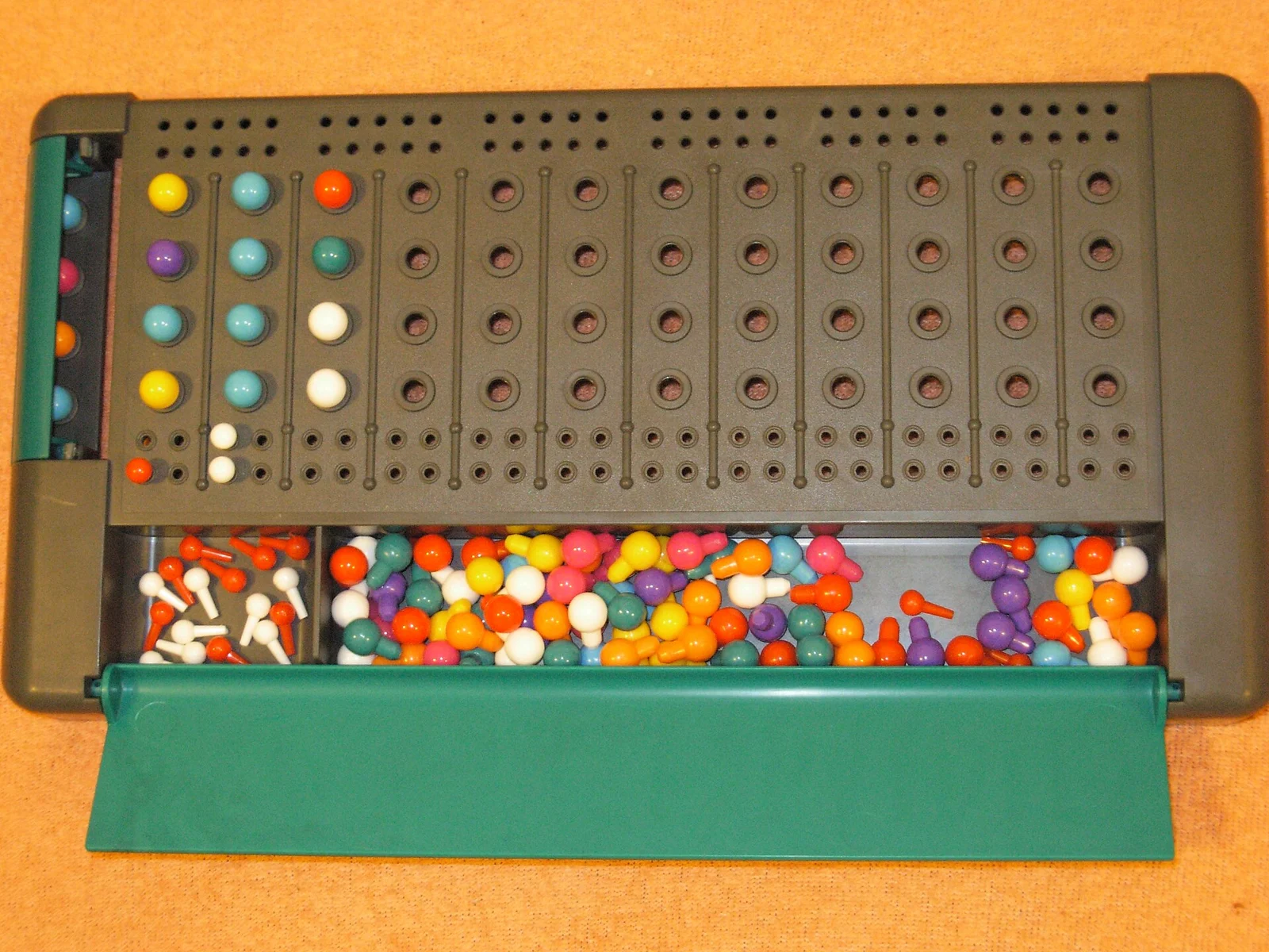 Aufgeklapptes Logikspielbrett mit zwölf Reihen und farbigen Steckkugeln sowie Vorratsfach.