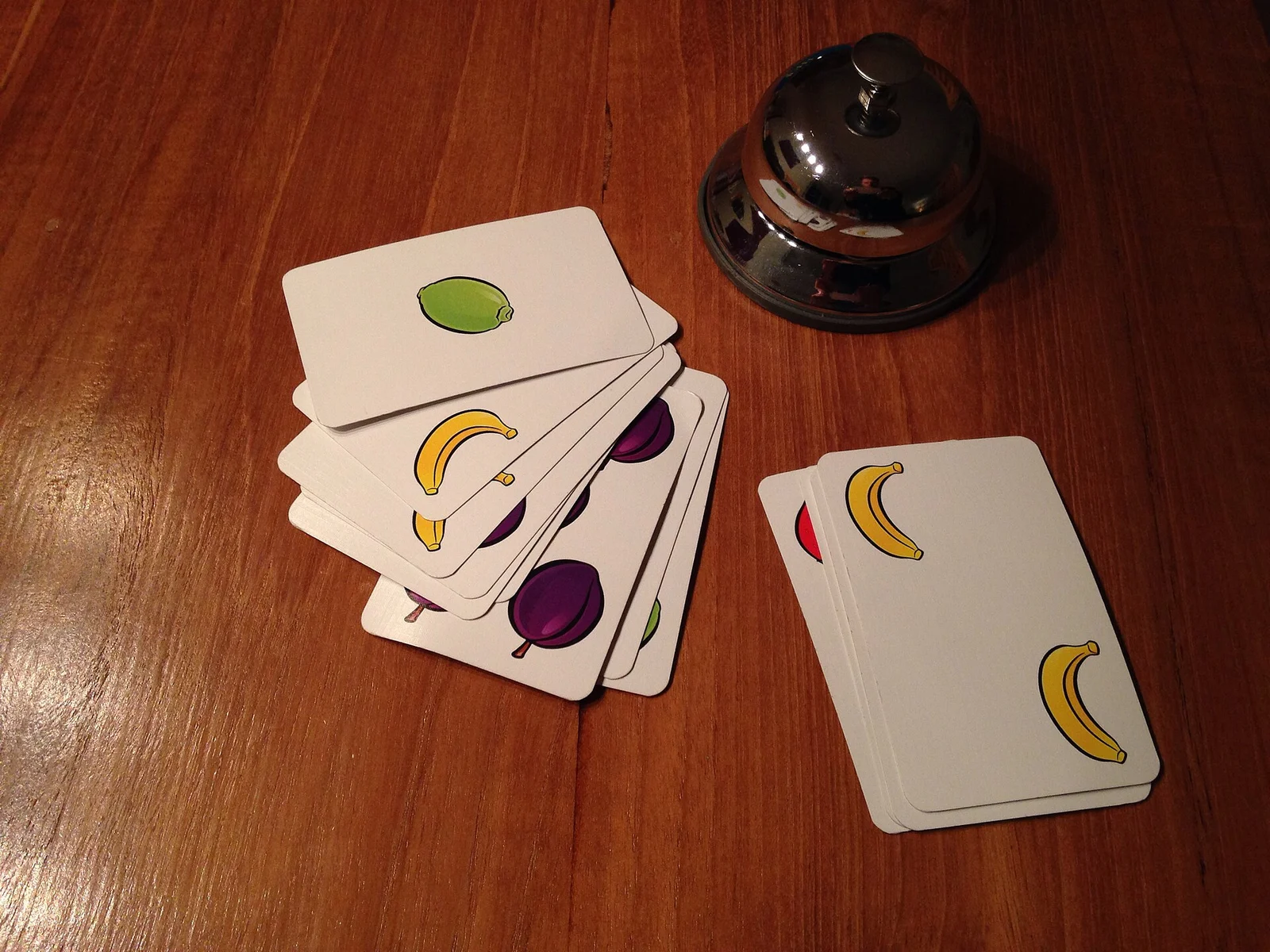 Spielkarten mit Obstmotiven und eine silberne Tischglocke auf einem Holztisch.