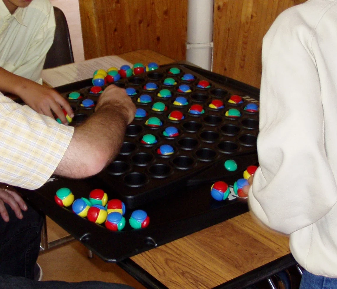 Zwei Personen setzen bunte vierfarbige Kugeln auf ein großes schwarzes Rolit-Spielbrett, nur Hände und Oberkörper sichtbar.
